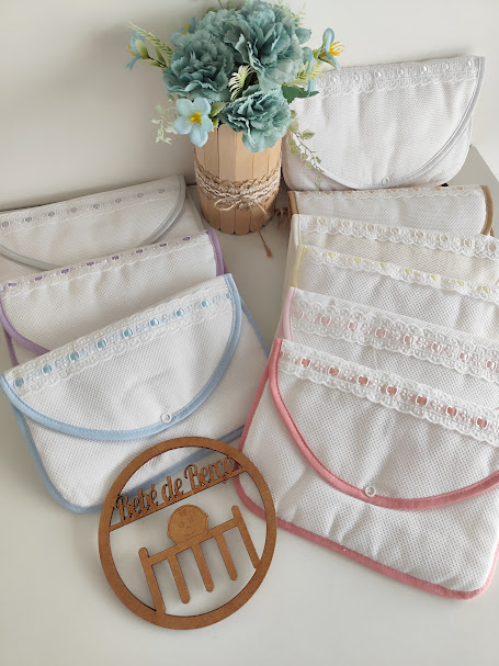 Capas de tecido branco com bordas coloridas e decoração em renda, vaso com flores azuis e círculo de madeira com texto