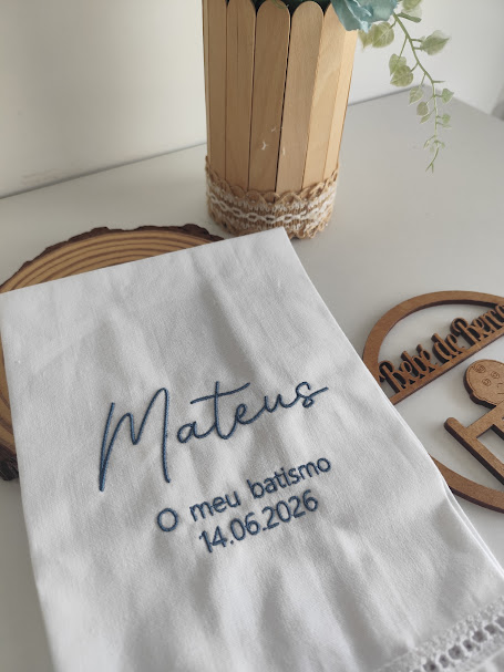 Toalha branca bordada com nome Mateus e data de batismo, perto de vaso decorativo