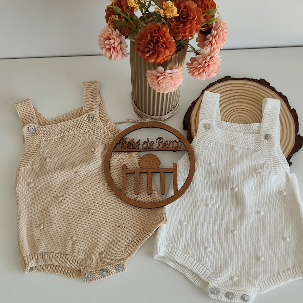 Dois macacões de bebé bege e branco em malha com pompons e botões, vaso de flores secs laranja/rosa e placa redonda de madeira com texto