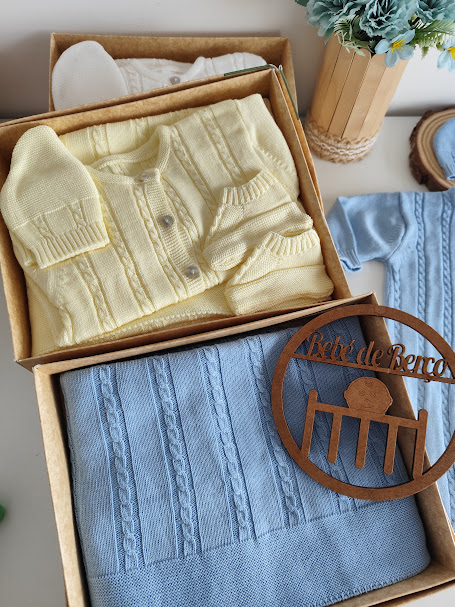 Conjunto de roupa de bebé em tricô amarelo claro e azul com manta, gorro, casaco e meias, em caixas de cartão, com planta e placa de madeira Bebé de Berço