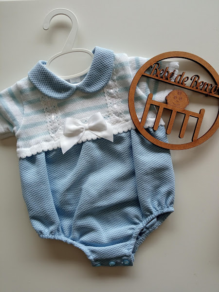 Body de bebé azul claro e branco com laço e renda, pendurado em cabide branco ao lado de placa de madeira com texto