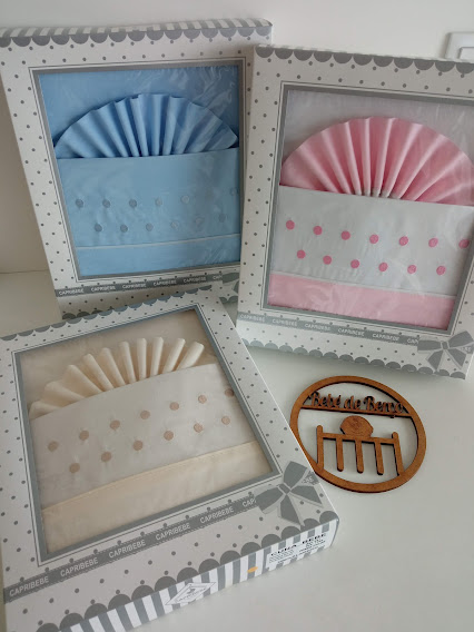 Conjuntos de roupa de cama para bebé em caixas de presente nas cores azul, rosa e creme com padrão de bolinhas bordadas, junto a ornamento de madeira redondo