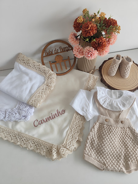 Conjunto de roupa de bebé com corpo branco e jardineiras bege de malha, manta bordada, sapatinhos e flores decorativas.
