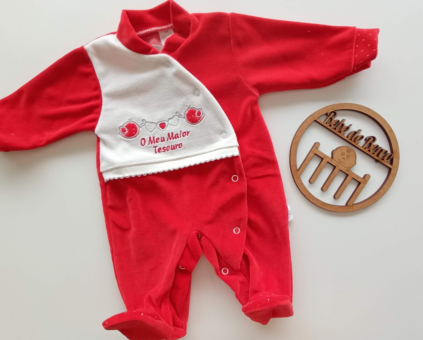 Macacão vermelho de bebé com manta branca bordada e objeto redondo de madeira