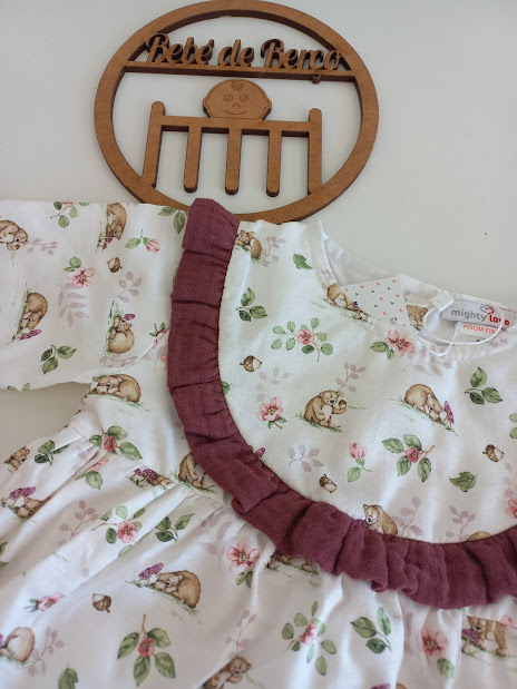 Vestido de bebé branco com estampado de animais e flores, detalhe roxo e etiqueta 'mighty L', pendurado junto a decoração de madeira