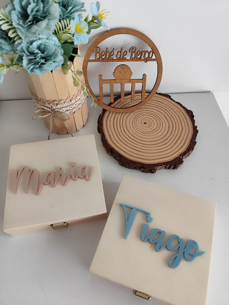 dois estojos brancos com nomes Maria e Tiago, suporte de madeira com texto Bebé de Romo, vaso com flores artificiais