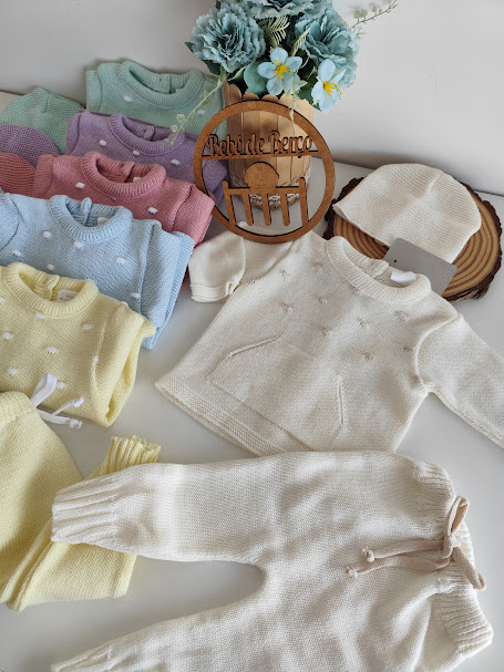 Conjunto de roupa de bebê branca em malha com outras roupas coloridas ao redor e flores azuis