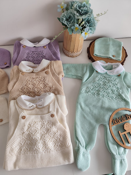 Conjunto de roupa de bebé em malha com padrões e botões, em várias cores, sobre mesa branca com flores e placa de madeira.