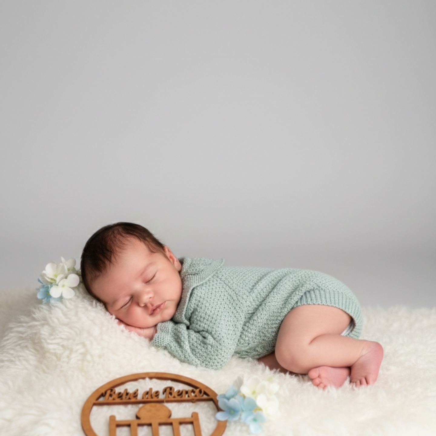 Bebé a dormir vestido de malha cinza-azulada sobre superfície macia branca com flores e placa de madeira Bébé de Berço.