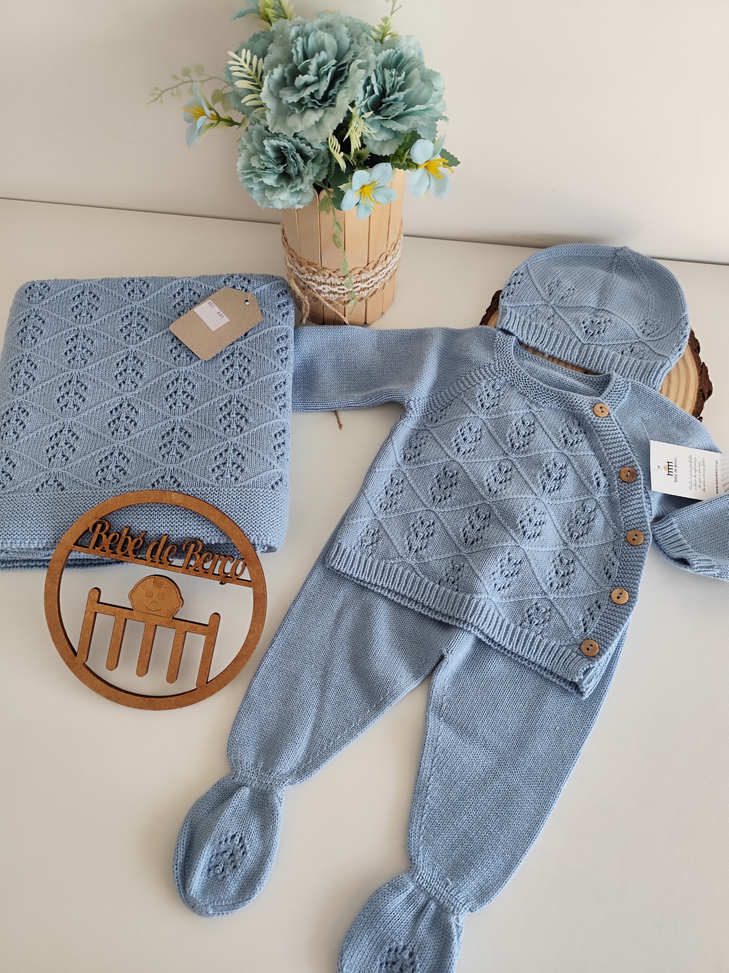 Conjunto de bebé azul com casaquinho, calças, gorro e manta com padrão em losangos sobre mesa branca com flores azuis