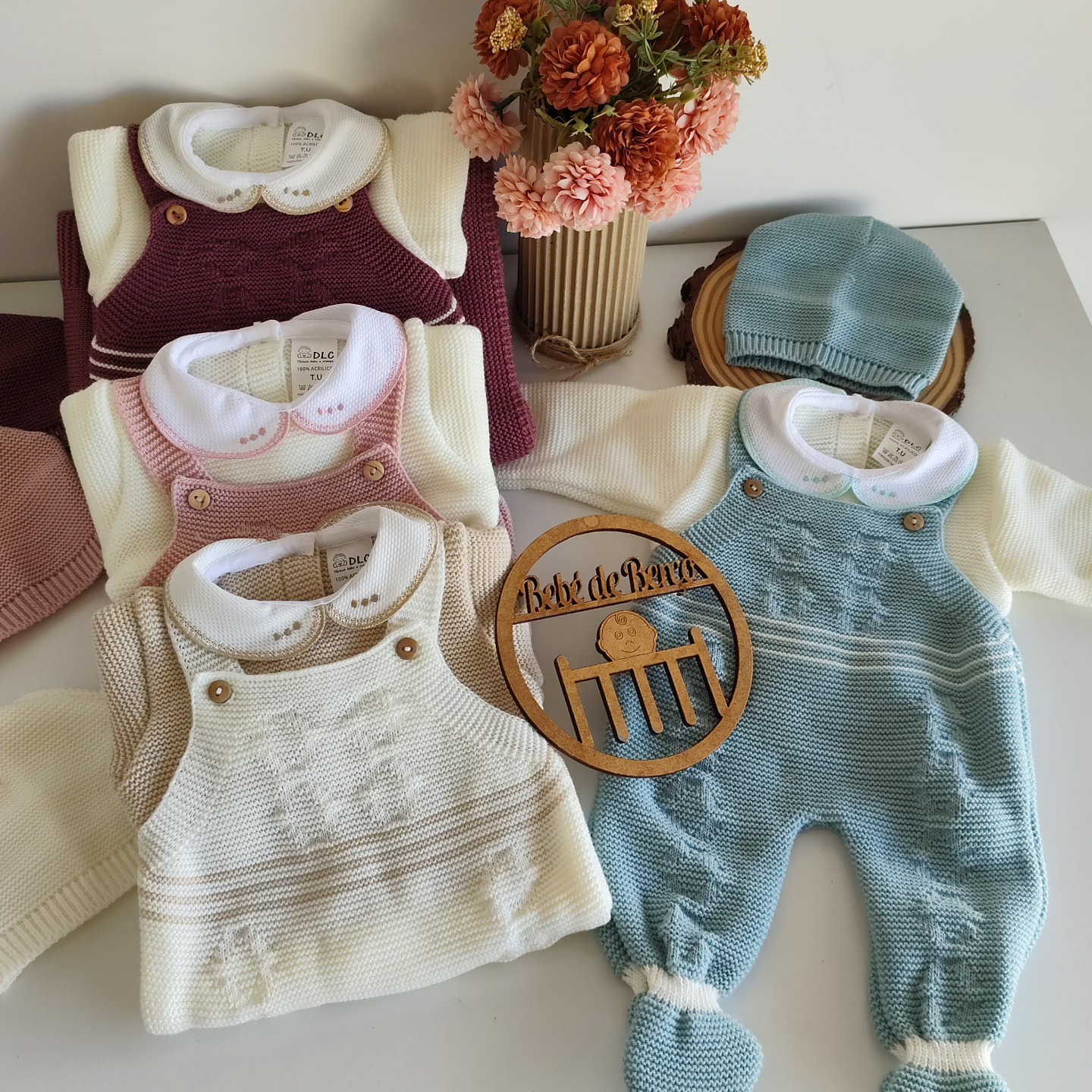 Conjunto de roupa de bebé em malha de várias cores com camisa branca, gorro azul, flores artificiais e placa de madeira com texto.