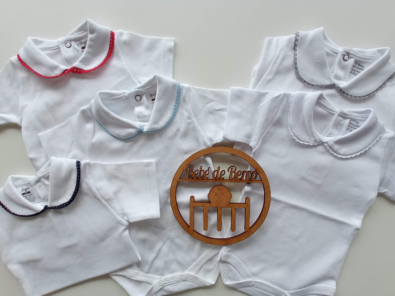 Cinco bodies brancos para bebés com golas de cores diferentes e placa de madeira com texto