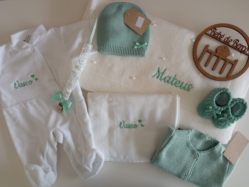 Conjunto de roupa de bebé branco e verde com nomes bordados e acessórios dispostos sobre superfície clara