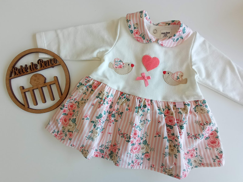 Vestido de bebé branco com saia floral rosa e aplicações de coração e passarinhos, ao lado círculo de madeira com texto Rita de Rato.