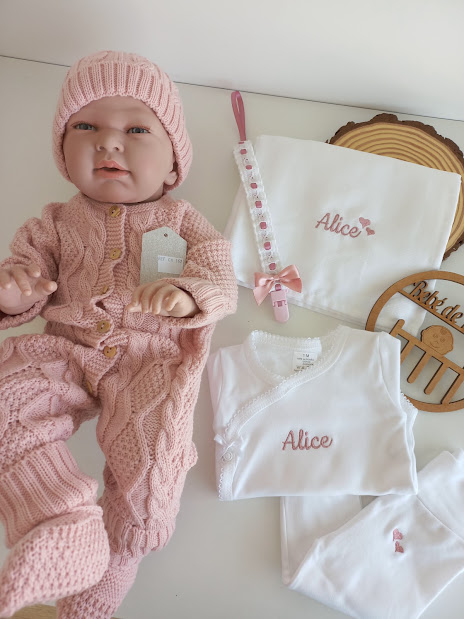 Boneca bebé vestida de malha rosa com conjunto branco bordado com nome Alice e chupeta com fita rosa