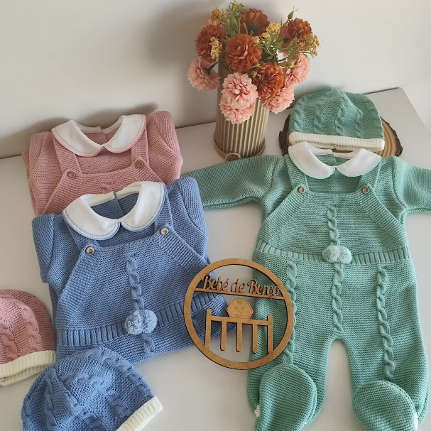 Conjunto de roupa de bebé em tricô colorida com gola branca e gorros, em cima de mesa com flores e placa de madeira