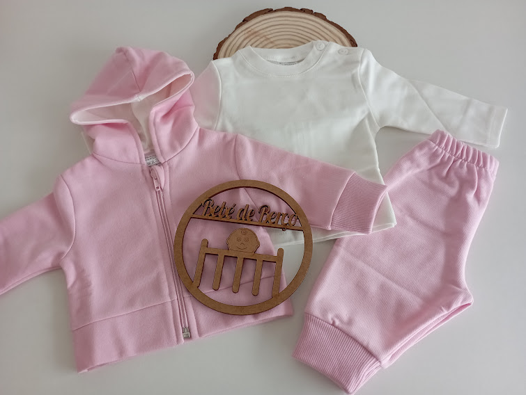 Conjunto de roupa para bebé com casaco, camisa e calças em rosa e branco sobre fundo branco com suporte de madeira decorativo