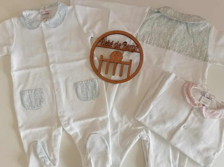 Três macacões de bebé brancos com detalhes azuis e rosas e placa de madeira com texto