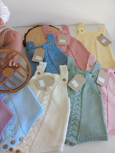 Vários macacões de bebê em cores pastel, feitos de malha com botões de madeira, dispostos sobre uma mesa branca