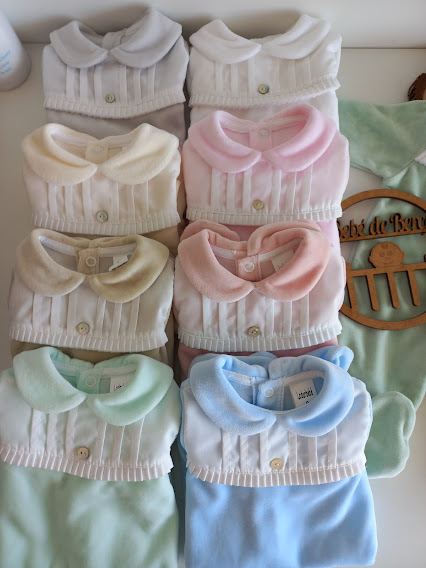 roupinhas de bebé dobradas em várias cores com gola e botões, junto a peça verde com símbolo em madeira sobre superfície branca