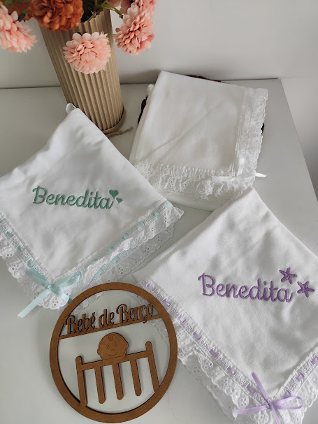 Três panos brancos bordados com o nome Benedita e um círculo de madeira decorativo num fundo branco com vaso de flores
