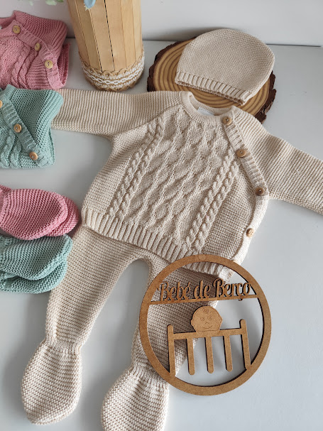 Conjunto de bebé em malha creme com camisola, calças com pé e gorro, com placas de madeira e outros conjuntos coloridos ao lado.