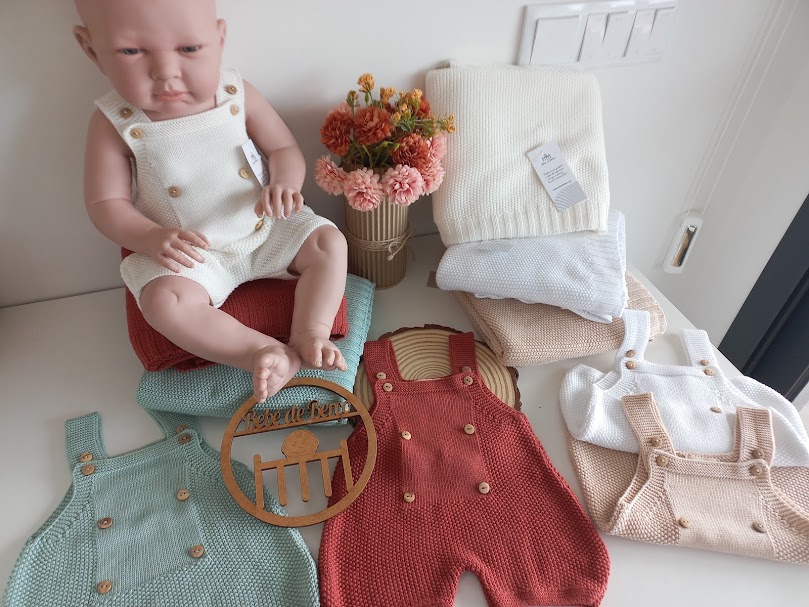 Boneca bebé com macacão de malha branco e vários macacões de malha coloridos com botões de madeira num fundo branco
