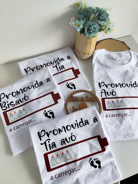Conjunto de t-shirts brancas com texto em português e gráficos de mamadeiras e pegadas, ao lado de vaso com flores azuis.