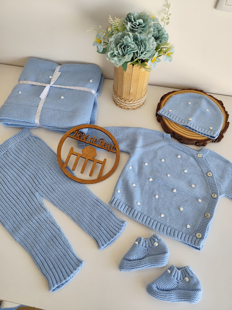 Conjunto de roupa azul claro para bebé com pompons brancos, placa de madeira e flores artificiais em vaso.