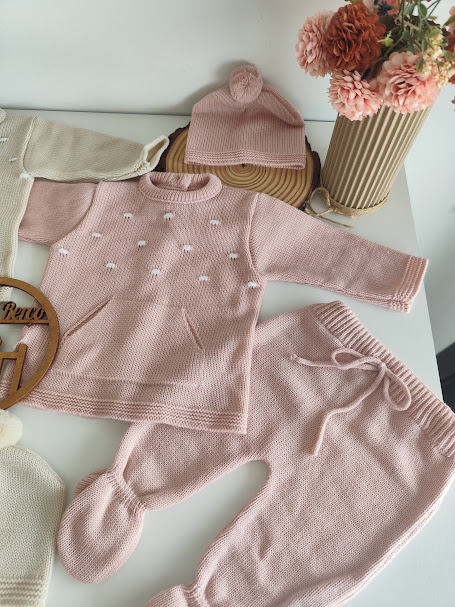 Conjunto de roupa bebé em malha rosa claro com camisola, calças e chapéu com pompom ao lado de vaso com flores