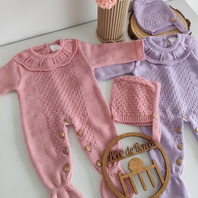 Macacões de bebé rosa e lilás de malha com botões e gorros, decorados com flores, placa de madeira na mesa