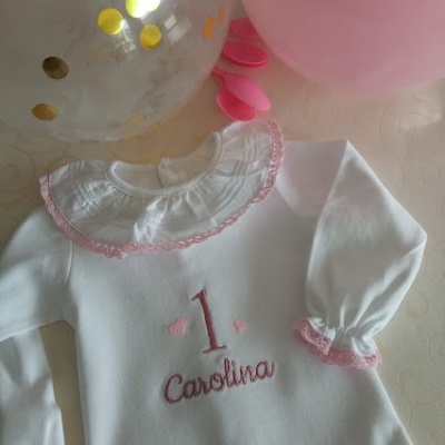 Macacão branco com bordado '1 Carolina' e gola com renda rosa, rodeado por balões rosa e transparentes