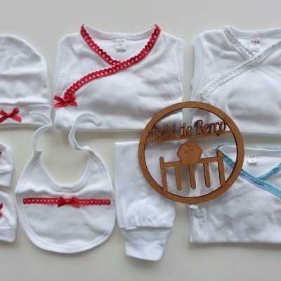 Conjunto branco de roupa de bebé com detalhes vermelhos e azuis e acessório de madeira redondo