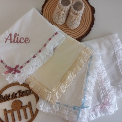 Quatro toalhas ou fraldas de pano com bordados em renda e laços coloridos, umas sapatilhas bege e suporte de madeira.