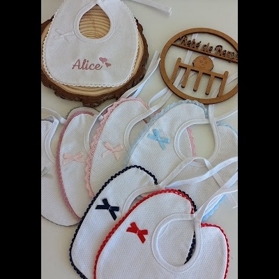 Babetes brancos bordados com laços coloridos e um com o nome Alice, ao lado de enfeite de madeira