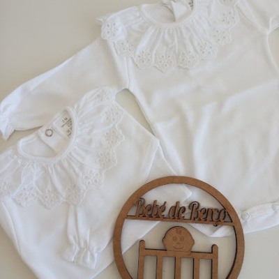 Dois bodies brancos de bebé com golas rendadas e placa em madeira com texto 'Bebé de Berço'