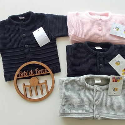 Quatro casacos de bebé em malha, azul-marinho, rosa, cinzento e preto, com botões e etiquetas, sobre fundo claro, junto a placa de madeira 'Bébé de Berço'.