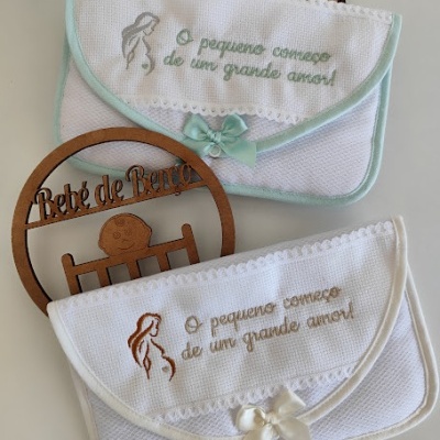 Duas bolsas de pano branco bordadas com laços e mensagem em português, sobre uma superfície clara com ornamento de madeira
