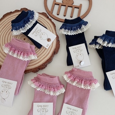 Meias de bebé rosa e azul com folhos rendados e etiquetas com texto sobre superfície branca e base de madeira