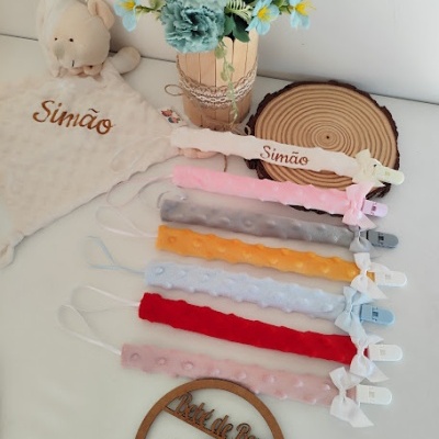 Fitas porta-chupetas coloridas e texturizadas com molas e laços, decoradas num fundo branco com peluche e flores artificiais.