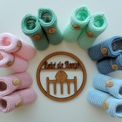 Vários sapatinhos de bebé em crochet de cores pastel dispostos em círculo em torno de um ornamento de madeira com texto.
