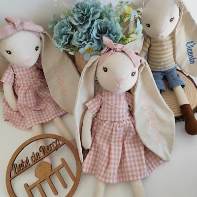 Três bonecos de coelho de tecido com roupas e orelhas compridas bordadas, vaso de flores azuis e placa de madeira com texto Petite de Perce.