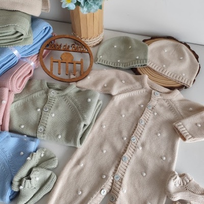 Roupas de bebé em malha creme, verde e azul com bolinhas, placa de madeira e vaso de flores