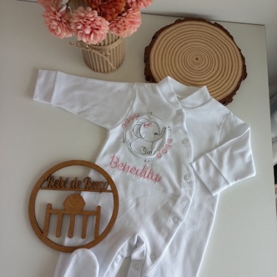 Macacão branco de bebé com bordado de elefante e nome Benedita, em superfície branca com placa de madeira e flores.