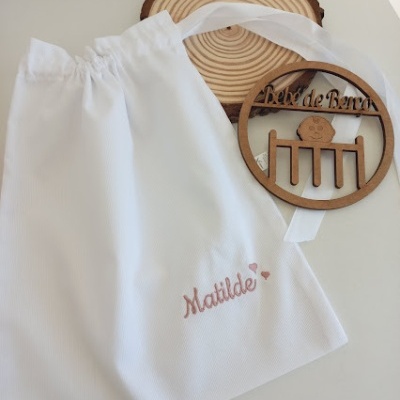 Saco branco com nome Matilde bordado ao lado de medalhão de madeira com texto e desenho de berço