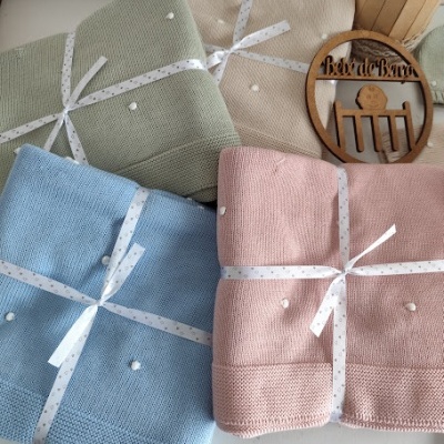 Mantinhas de malha azul, rosa, verde e bege com pompons e fita decorativa em superfície branca