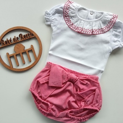 Conjunto de roupa de bebé em branco e cor-de-rosa com body e cueiros, junto a placa redonda de madeira com texto