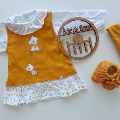 Conjunto de roupa de bebé com vestido amarelo mostarda, camisa branca, meias e sapatinhos amarelos, com placa de madeira Bebé de Pernas.