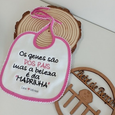Babete branco com bordas cor de rosa e texto bordado, colocado sobre uma base redonda de madeira e adereço de madeira com texto