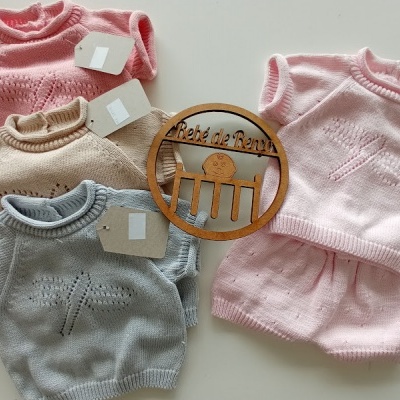 Quatro conjuntos de roupa de bebé em malha colorida com padrão rendado e etiquetas, sobre fundo branco com objeto decorativo de madeira